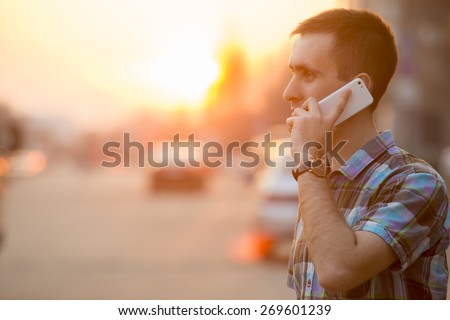 Young man holding mobile phone, using smartphone, making a call, talking on the phone, standing on sunny street with transport traffic on the background