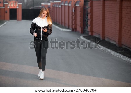 Beautiful sporty girl in sportswear walking in earphones during sport workout in the morning street holding device, looking at gadget, using app