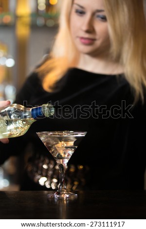 Beautiful blond bartender girl serving alcohol drink at bar counter, holding bottle in hand, pouring drink in cocktail glass, focus on beverage