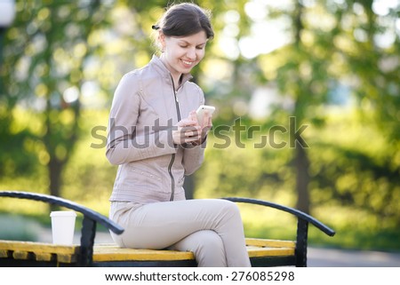 Portrait of happy smiling caucasian woman in park siting on the bench with takeaway drink, holding smartphone, using app, messaging, looking at screen
