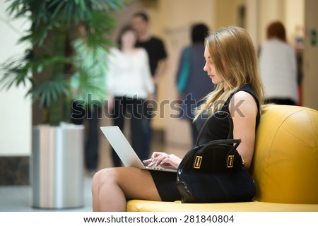 Young female sitting on yellow coach working on laptop in public wifi area, typing, people passing by on the background