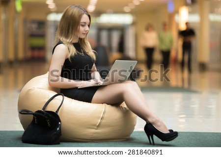 Young female sitting on beanbag working on laptop in public wifi area, typing, people passing by on the background