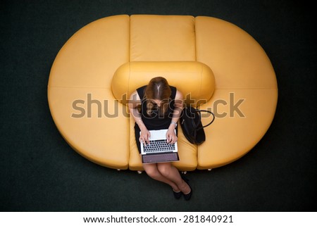 Young female sitting on yellow coach working on laptop in public wifi area, typing, top view