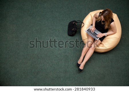 Young female sitting on bean bag working on laptop in public wifi area, typing, top view, copy space