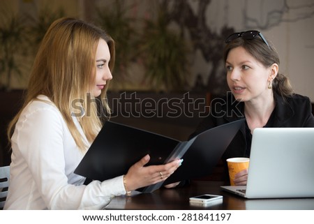 Two young caucasian women sitting at the table in cafe beside laptop, having discussion, blond girl holding open document folder, listening