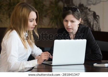 Two young caucasian office women meeting, having serious discussion, sitting at the table, using laptop, looking at screen