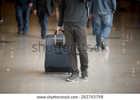 Young man with suitcase walking in modern airport terminal with crowd on background, wearing casual style clothes, close up