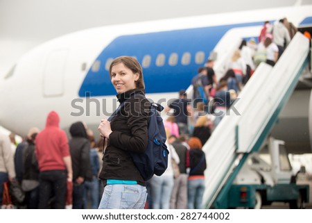 Young Caucasian happy smiling woman passenger in 20s travelling with backpack, boarding airplane, looking at camera, people climbing air stairs on background