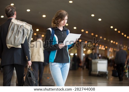Portrait of young woman in 20s with backpack standing in airport terminal, with printed e-tickets, using cell phone to check in, blurred crowd of travelling people on the background