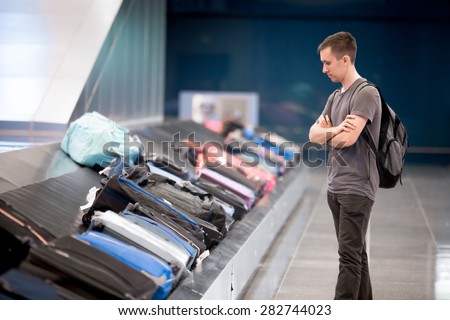 Young handsome man passenger in his 20s with carry-on backpack waiting at conveyor belt to pick his luggage in arrivals lounge of airport terminal building