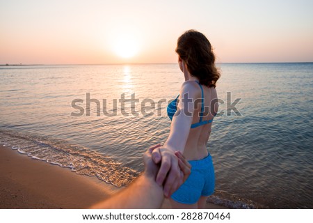Young woman in blue swimwear leading by hand her lover, at the sea coast in sunlight, follow me concept