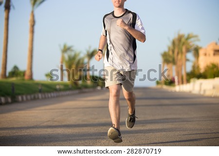 Healthy lifestyle: close up of sporty young man torso and legs in running shoes, jogging on the road on the sunny summer street in tropics, palm trees on the background