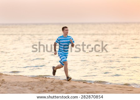 Healthy lifestyle: handsome cheerful young man working out outside, running fast on the sand coast at dusk on sunset or sunrise