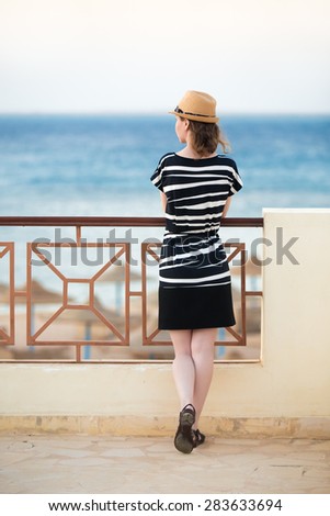 Young woman in hat and cute summer dress standing in feminine carefree pose at the balcony with beautiful sea scenery, enjoying landscape, back view