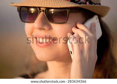 Portrait of young Caucasian happy smiling woman in cute straw hat and sunglasses using cellphone, making call in sunlight, close up