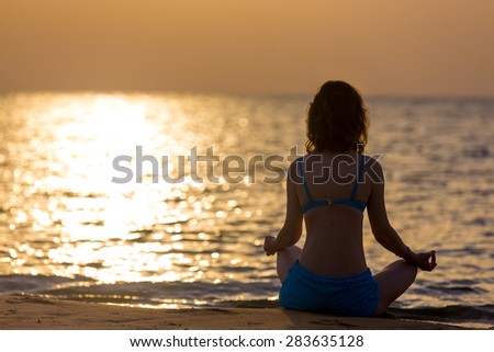 Serene young woman practicing yoga, Sitting in Easy (Decent, Pleasant Pose), Sukhasana, meditating, relaxing on the sea coast at sunset or sunrise, copy space, back view