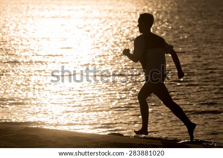 Healthy lifestyle: silhouette of sporty young man working out on the beach, running fast on the sand shore against the sea lighted with golden sun