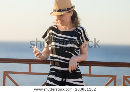 Young happy smiling woman in cute straw hat and casual summer dress leaning on railing on sunny street of sea town, holding cellphone, looking at screen, using app, messaging
