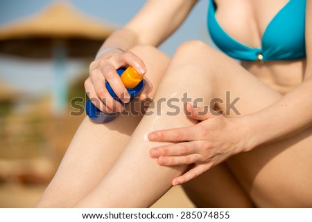 Young woman in swimsuit holding bottle of sunscreen lotion, applying sunblock cream on legs before tanning on sunny southern sand beach with straw umbrellas, close up