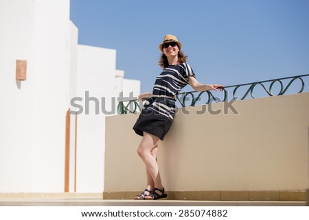Portrait of young happy smiling woman in hat and cute summer dress standing at sunny summer street with white stucco buildings, enjoying sunshine