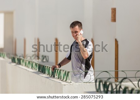 Portrait of young man standing on balcony of white stucco building in bright sunlight, making call, talking on mobile phone, using cellphone