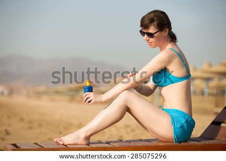 Young smiling woman in blue swimwear on sunny beach holding bottle of suntan lotion, applying sun cream on her arm before tanning