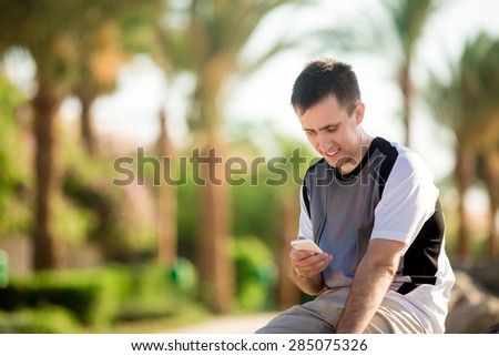 Young handsome happy smiling man sitting in relaxed pose on southern sunny street or park, holding cellphone, making call, messaging, using app, copy space