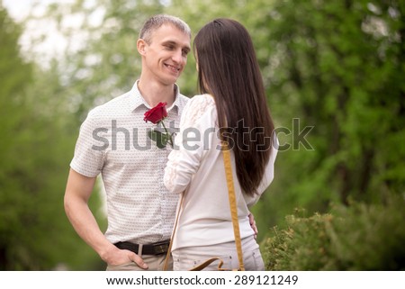 Couple of lovers meeting on a date in park, looking each other in eyes, smiling young man giving red rose to his brunet girlfriend