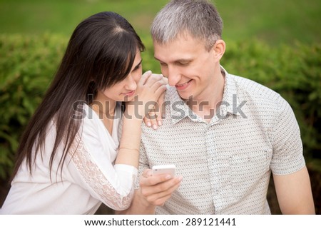 Young attractive happy smiling lovers couple in love sitting on park bench, dating, hugging, holding cell phone, looking at screen together, using app, absorbed in phone communication