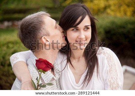Portrait of couple with red rose on date in park, young man kissing his girlfriend in her ear while she is sitting on his laps with happy playful smile