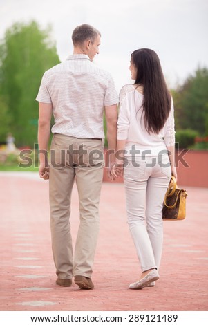 Couple of lovers, attractive young man and woman dating, walking holding hands in park, full length, back view