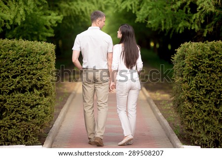 Couple in love walking in park holding hands, attractive young man and woman on date, looking at each other, talking, full length, back view