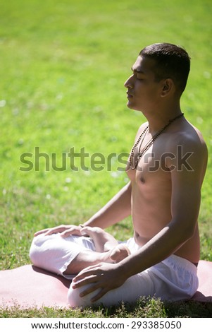 Serene Indian young man in white pants practicing yoga on green grass in park, doing Padmasana, sitting with closed eyes in Lotus Pose, breathing, meditating, relaxing, full length, vertical shot