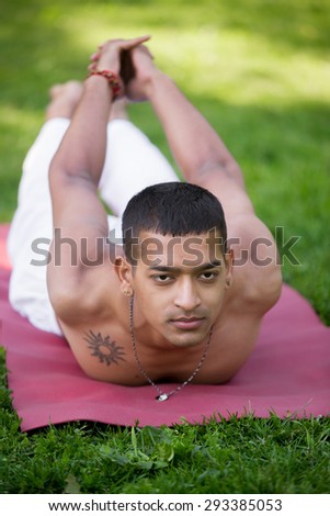 Indian young man with tattoo in white linen clothes practicing yoga, fitness, pilates in park, doing back extension exercise, shalabhasana, Locust Pose, Double Leg Kicks, full length, front view