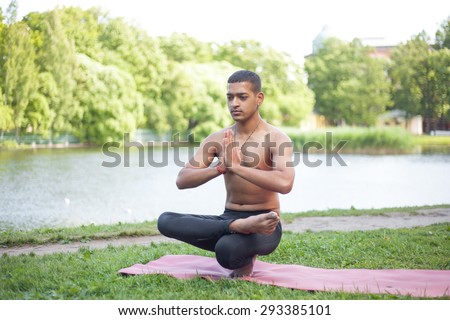 Sporty handsome Indian young man working out on mat on lake in park, sitting in squat, doing Half Lotus Toe Balance, Padangustha Padma Utkatasana, hands in Namaste, full length