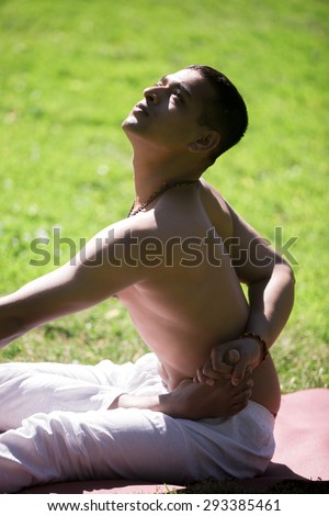 Serene Indian young man in white pants practicing yoga, fitness or pilates on green grass in park, doing ardha baddha padma paschimottanasana, half bound lotus forward bend, close up