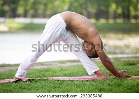 Profile of attractive Indian young man in white linen clothes practicing yoga, fitness, pilates on lake in park, standing in Pyramid Pose, Parsvottanasana, deep variation, full length