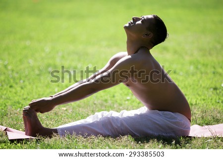 Serene Indian young man in white pants practicing yoga, fitness or pilates on green grass in park, doing seated Forward Bend pose, Paschimottanasana, stretching spine, full length