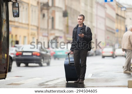 Young smiling handsome man in 20s with luggage bag standing on rainy city street with busy traffic transport using smartphone, waiting for public bus, traveling, wearing casual style clothes