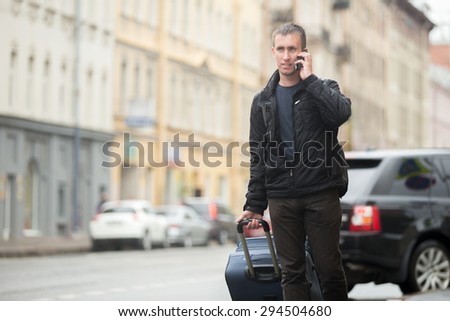 Young smiling handsome man with luggage bag walking on rainy city street with busy traffic holding cellphone, making call, talking on mobile phone, travelling, wearing casual clothes