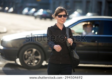 Young cheerful smiling woman with backpack crossing the road at pedestrian crosswalk, walking in sunny European city street in summer, blurred riding car on the background