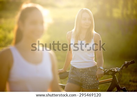 Two attractive girlfriends wearing casual white tank tops and jeans shorts standing with bikes in countryside park on bright sunny summer day, focus on beautiful blond young model
