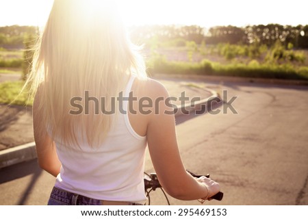 Beautiful teenage girl riding bike wearing casual white tank top and jeans shorts on park road on bright sunlit summer day, back view, copy space