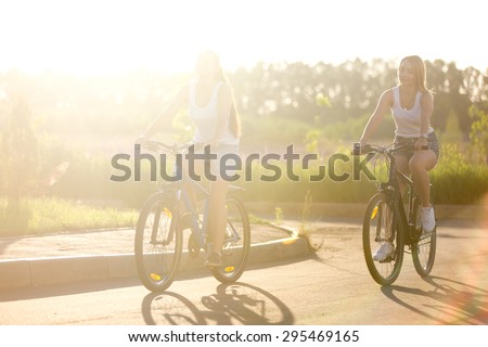 Two beautiful teenage girls riding bikes on park track wearing casual white tank tops and jeans shorts on bright sunny summer day, full length