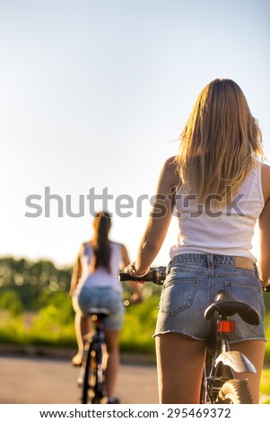 Back view of two sporty beautiful young women on bikes wearing casual white tank tops and jeans shorts on park road on bright sunny summer day
