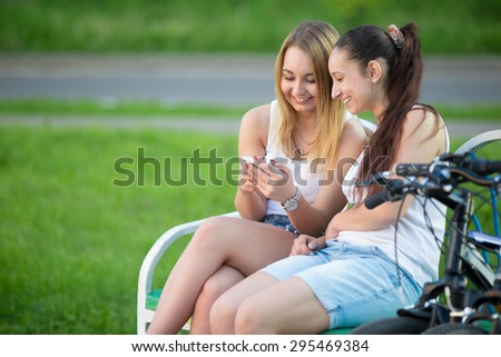 Portrait of two happy smiling beautiful young women friends wearing casual clothes sitting on park bench on summer day, having good time together, using app on smartphone, copy space
