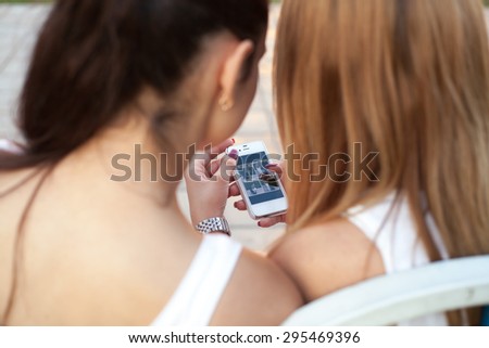 Two cute beautiful young women friends sitting on park bench on summer day, browsing pictures on smartphone, back view, focus on device screen