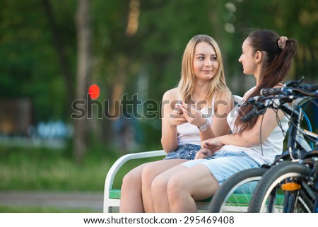 Two happy smiling beautiful young biker women friends wearing casual clothes sitting on park bench on summer day, chatting, having good time together, copy space