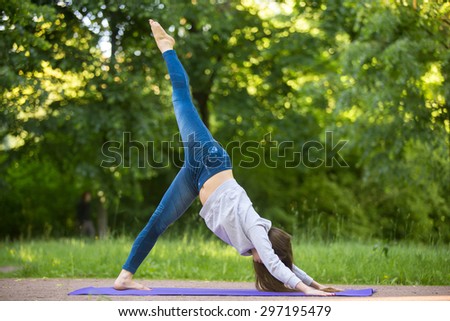 Profile of beautiful sporty young woman working out on blue mat on the street in park alley, doing eka pada adho mukha svanasana, one legged downward-facing dog pose, full length