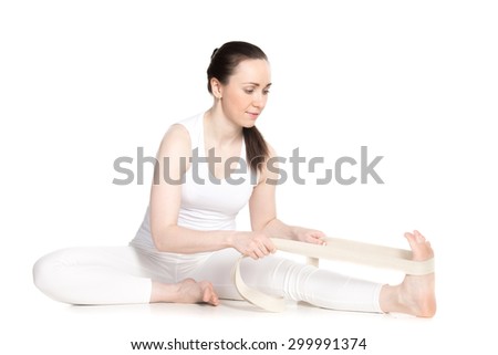 Sporty beautiful young beginning yoga student in white sportswear sitting in head to knee forward bend pose, doing Janu Sirsasana variation with strap, studio full length isolated shot